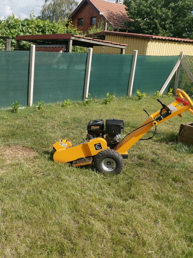 Sárga tuskómaró gép füves kertben, háttérben zöld árnyékoló kerítés és faház. A motoros berendezés fatuskók eltávolítására szolgál.